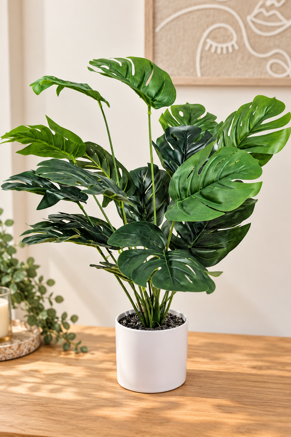 An artificial Monstera deliciosa plant with realistic green leaves in a white cylindrical pot, displayed on a wooden table in a warm, modern interior setting.