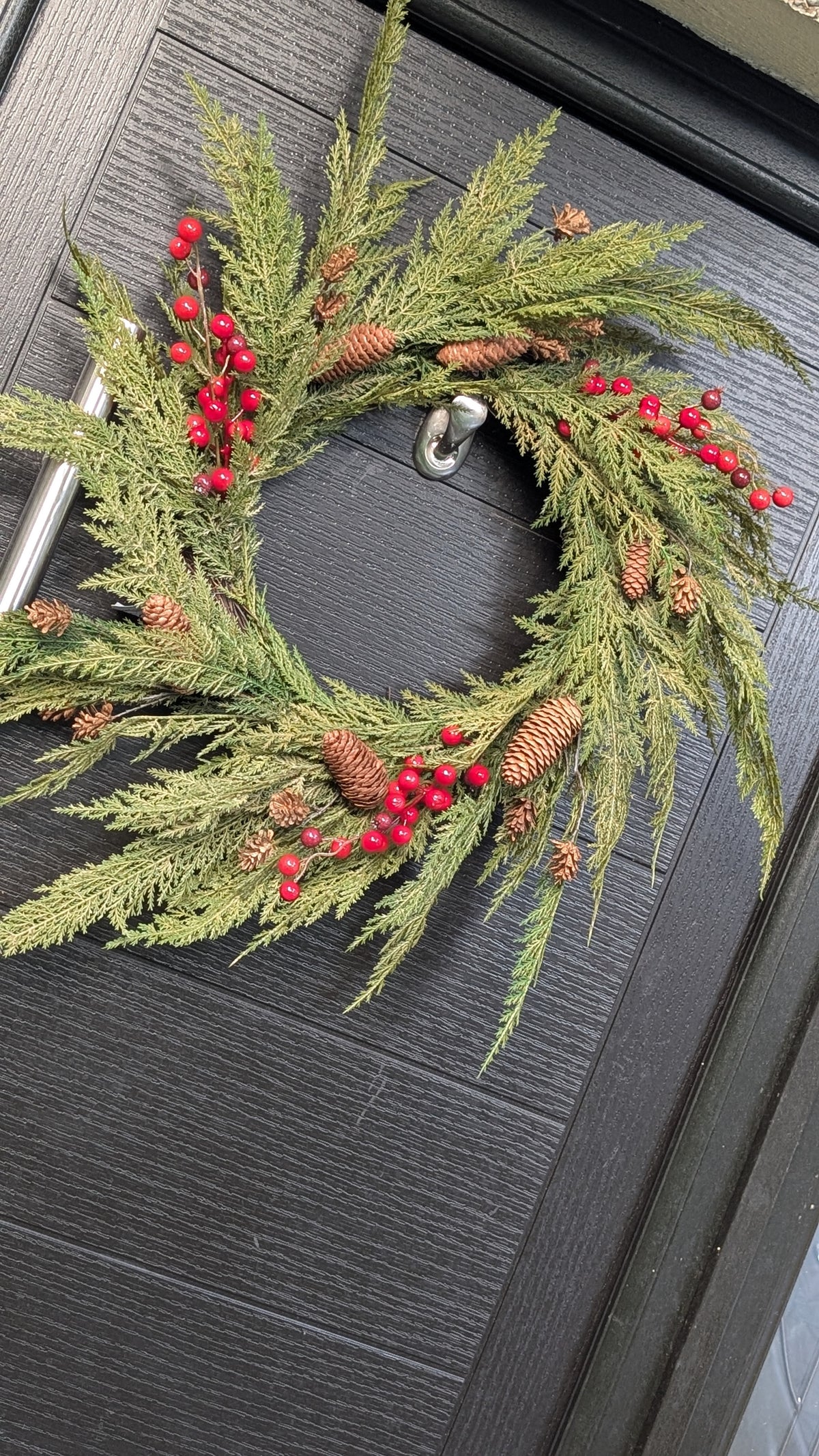 60cm ready-made Cupressus Christmas wreath with cascading green foliage, red berries and natural pinecones displayed on a dark front door.