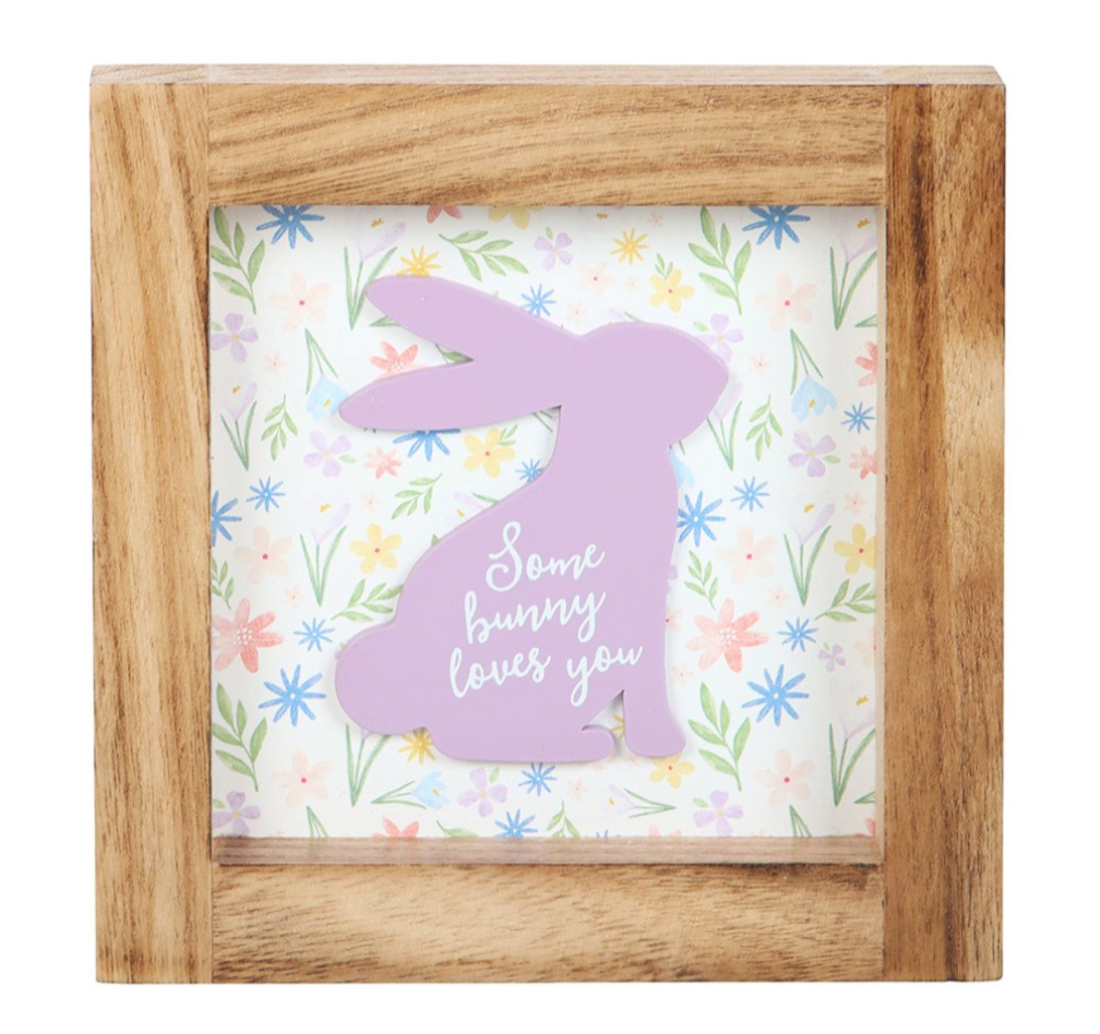 A square wooden shadow box sign featuring a pastel purple bunny silhouette with the phrase 'Some bunny loves you' in white script, set against a floral patterned background.