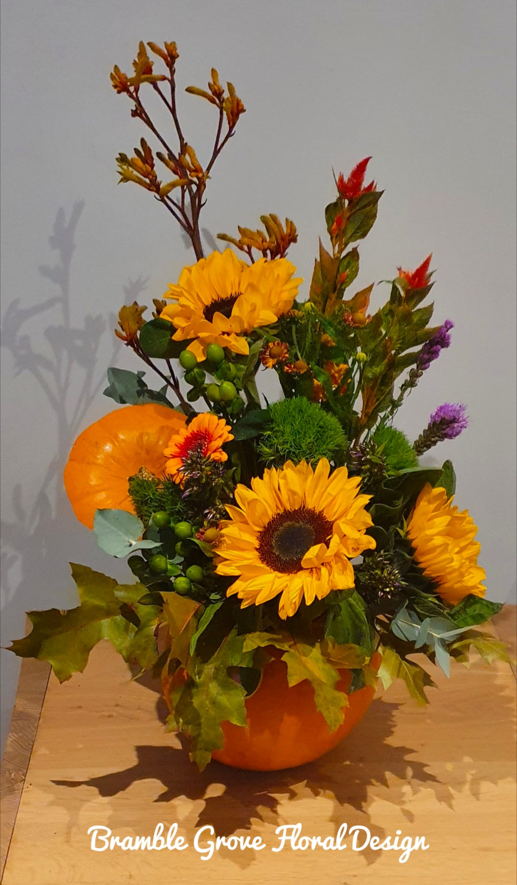 An autumn floral arrangement in a pumpkin-shaped vase, featuring large yellow sunflowers, orange pumpkins, green foliage, and small red and purple blossoms, displayed on a wooden surface against a plain gray wall.