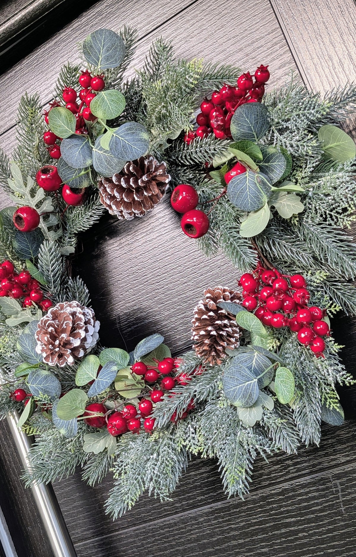 55cm traditional Christmas wreath featuring frosted evergreen foliage, red berries, snowy pinecones and eucalyptus-style leaves against a dark front door.