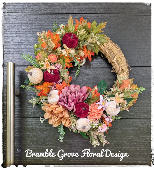 An autumn-themed floral wreath with faux straw, orange and pink flowers, burgundy roses, small pumpkins, and green leaves, displayed on a dark gray wood plank door. The text 'Bramble Grove Floral Design' is written at the bottom.