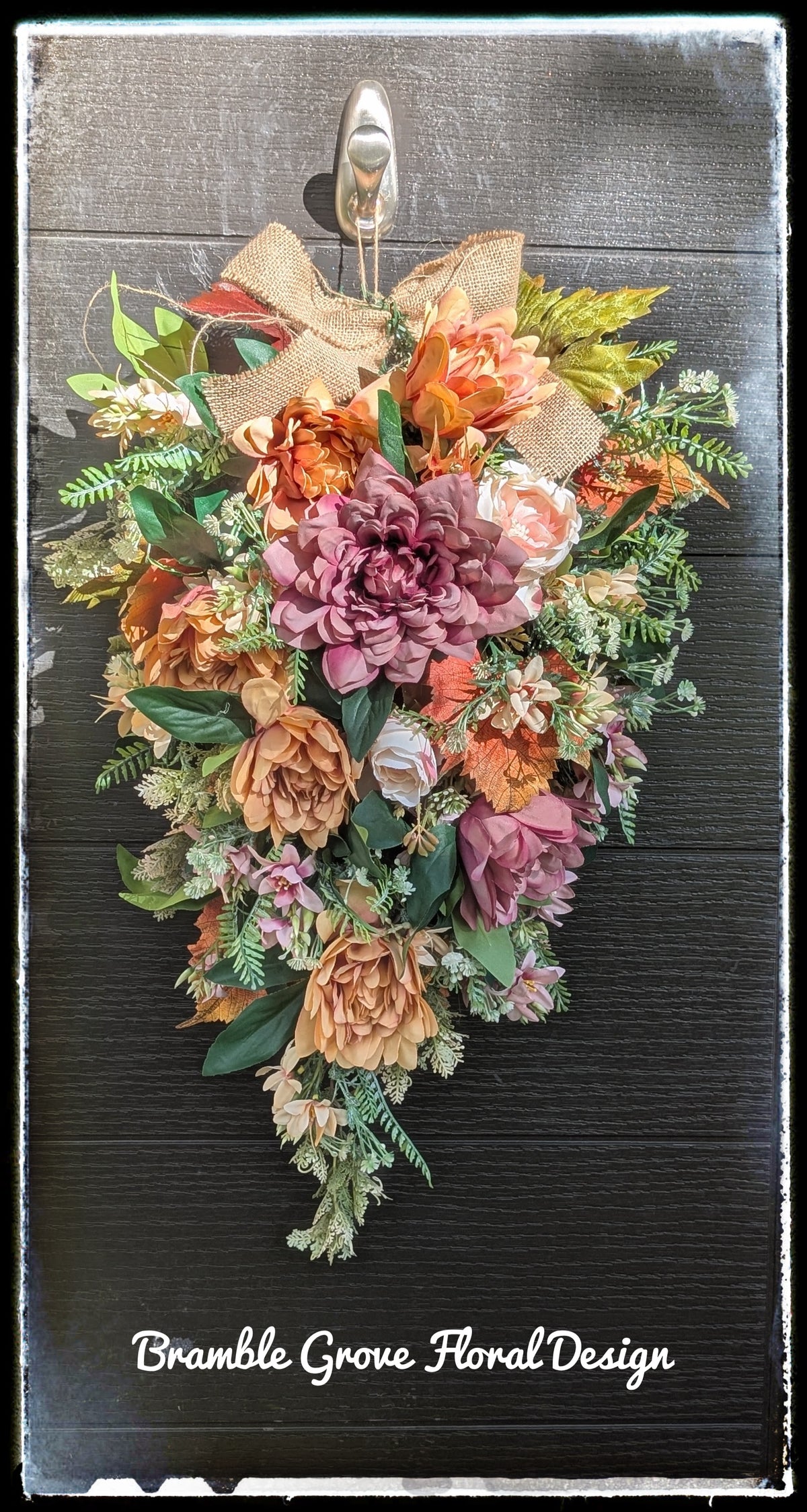A rustic heart-shaped floral wreath with faux flowers in shades of pink, orange, and beige, accented with green foliage and a burlap bow, hanging on a dark wood door. The text 'Bramble Grove Floral Design' is visible at the bottom.
