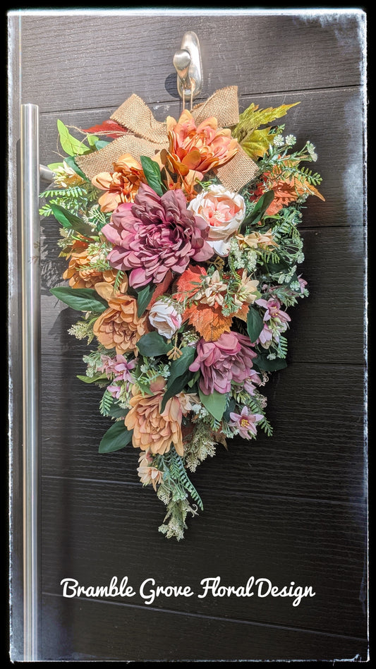 An autumn floral wreath with orange, pink, and peach artificial flowers, green leaves, and a tan burlap bow, hanging on a dark wood door. The text 'Bramble Grove Floral Design' is visible at the bottom.