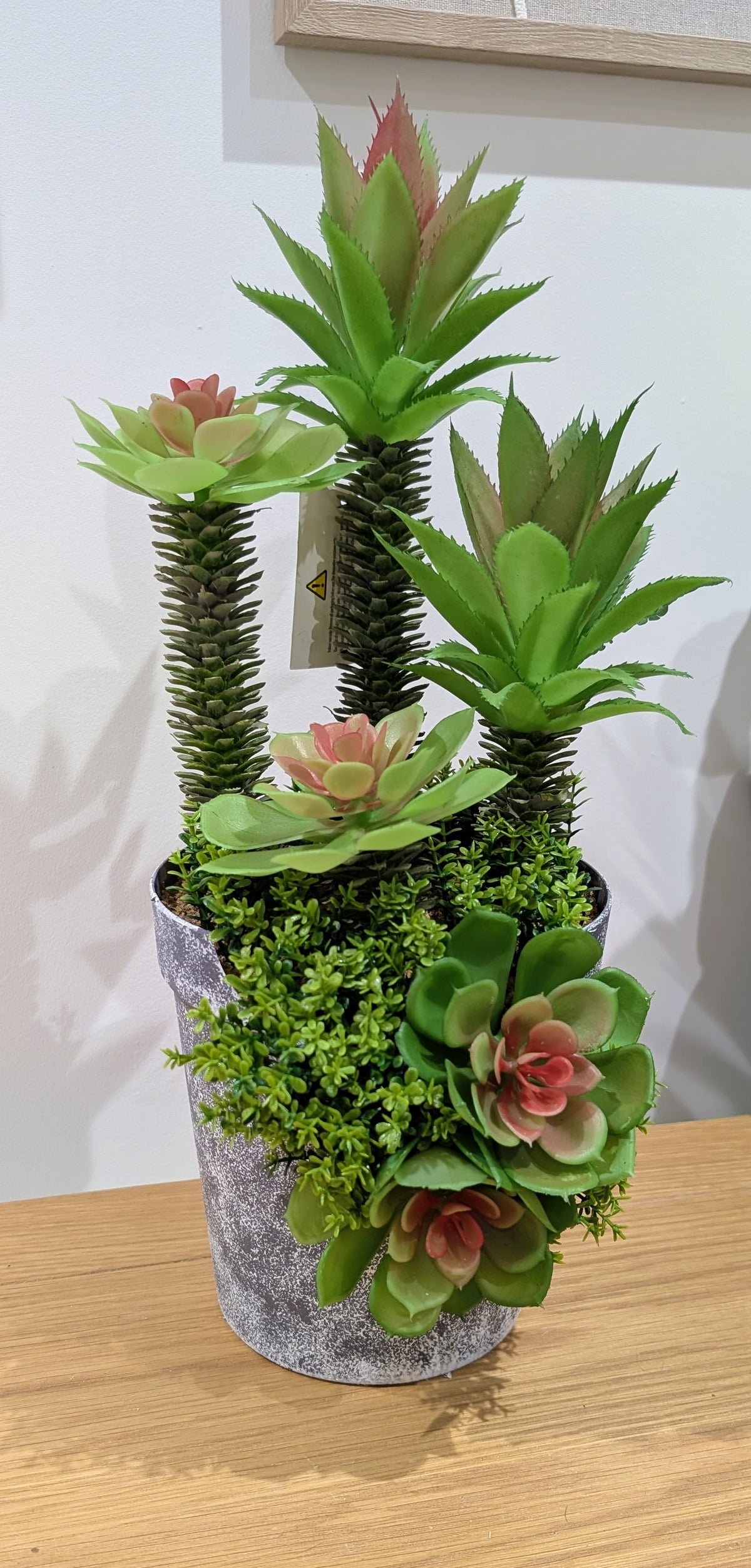 A potted artificial succulent and moss arrangement in a gray textured bucket-style planter, displayed on a wooden surface against a white wall.
