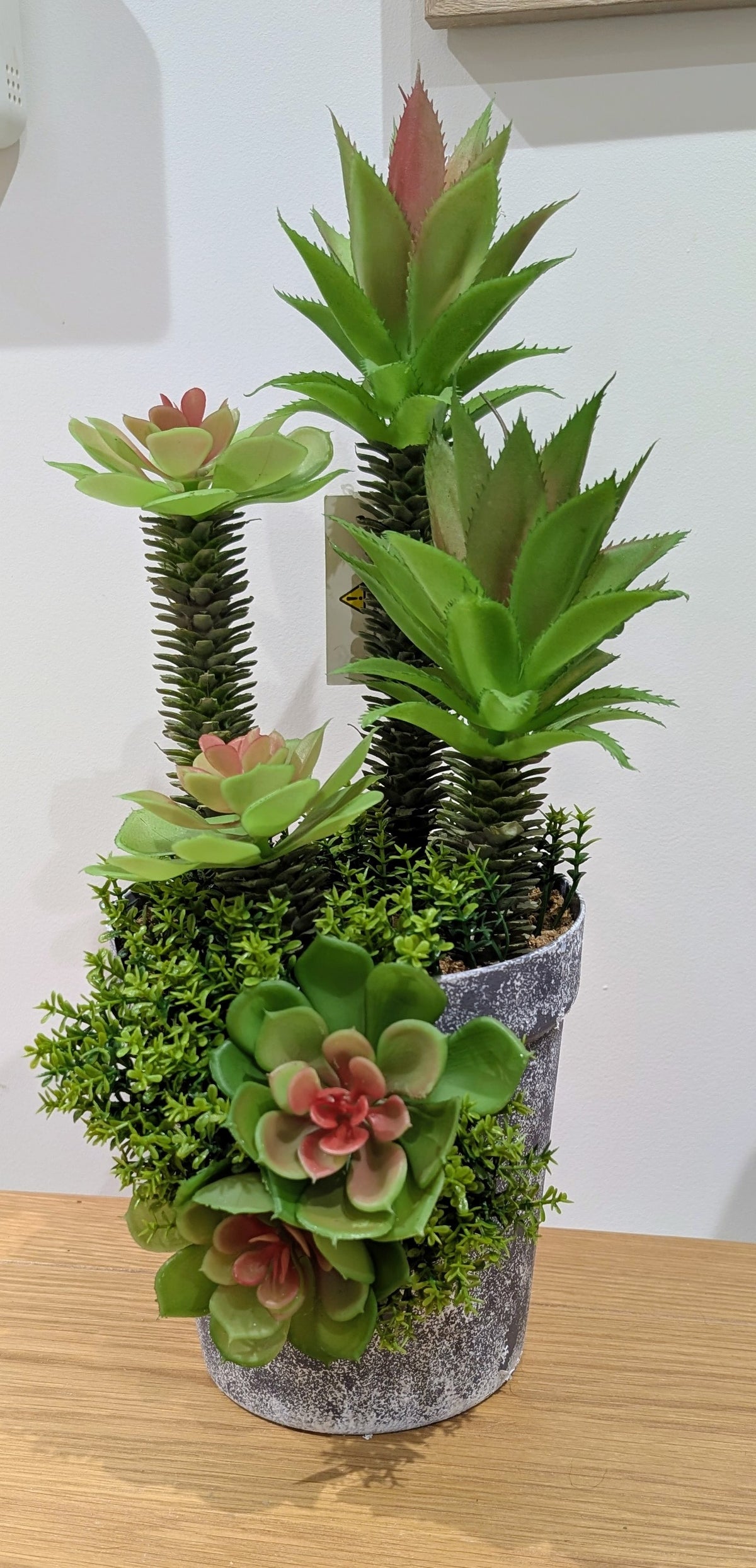 A potted artificial succulent and moss arrangement in a gray textured pot, displayed on a wooden surface against a plain indoor wall.