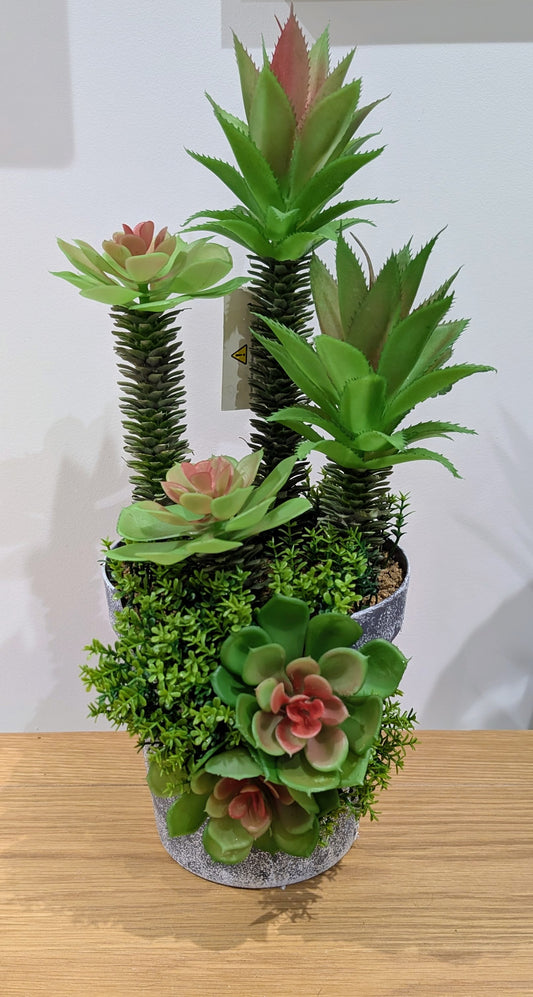 A potted artificial succulent and moss arrangement with multiple green faux plants and pinkish rosettes in a gray speckled pot, placed on a wooden surface against a plain light background.