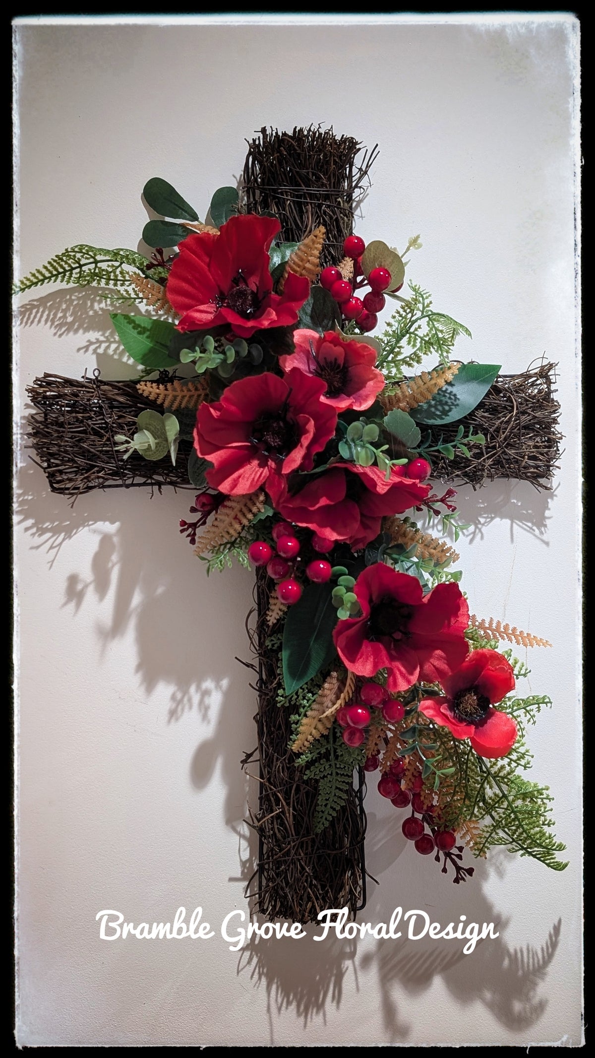 Rustic rattan Remembrance cross featuring cascading red artificial poppies, glossy red berries and mixed green foliage against a plain wall.