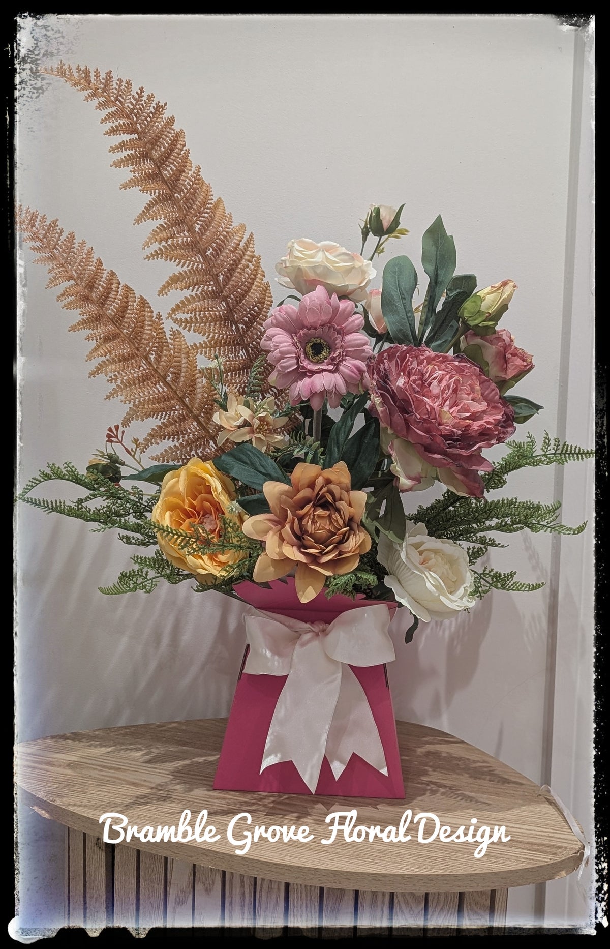A floral arrangement in a pink gift box with a white ribbon, displayed on a wooden surface against a light background. The bouquet includes pink, yellow, and peach flowers with green and tan foliage.