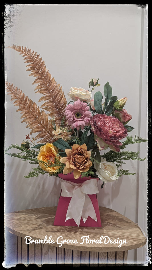 A floral arrangement in a pink gift box with a white bow, placed on a wooden surface against a light wall. The bouquet includes pink, peach, yellow, and white flowers with green foliage and a large tan fern frond.