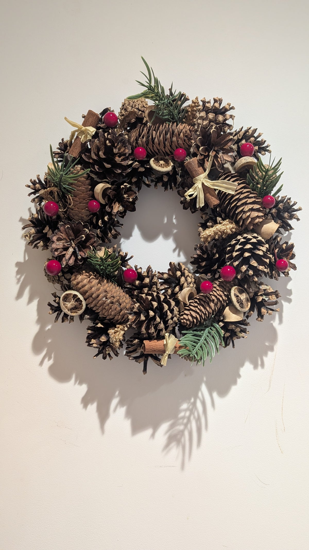 A rustic pinecone wreath decorated with natural brown pinecones, small green pine branches, bright red berries, and light wood accents, hanging against a plain off-white wall.