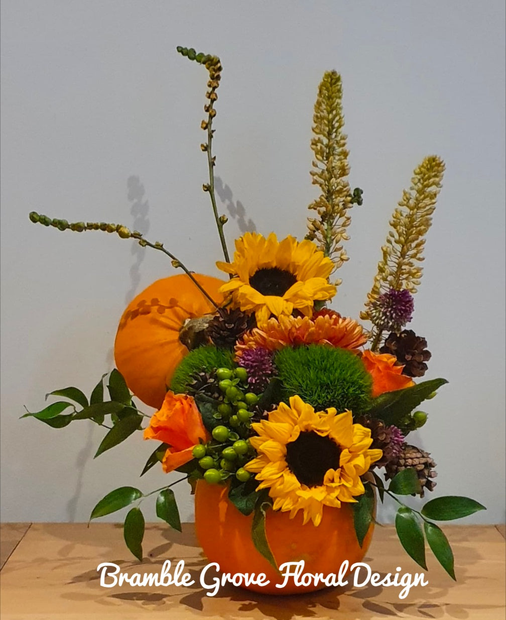 An autumn floral arrangement featuring sunflowers, orange mums, green mums, berries, and foliage in an orange pumpkin-shaped vase, with a small orange pumpkin behind it, set on a wooden surface against a plain light gray wall. The text 'Bramble Grove Floral Design' is overlaid at the bottom.