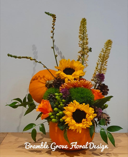 An autumn floral arrangement featuring sunflowers, orange mums, green mums, berries, and foliage in an orange pumpkin-shaped vase, with a small orange pumpkin behind it, set on a wooden surface against a plain light gray wall. The text 'Bramble Grove Floral Design' is overlaid at the bottom.