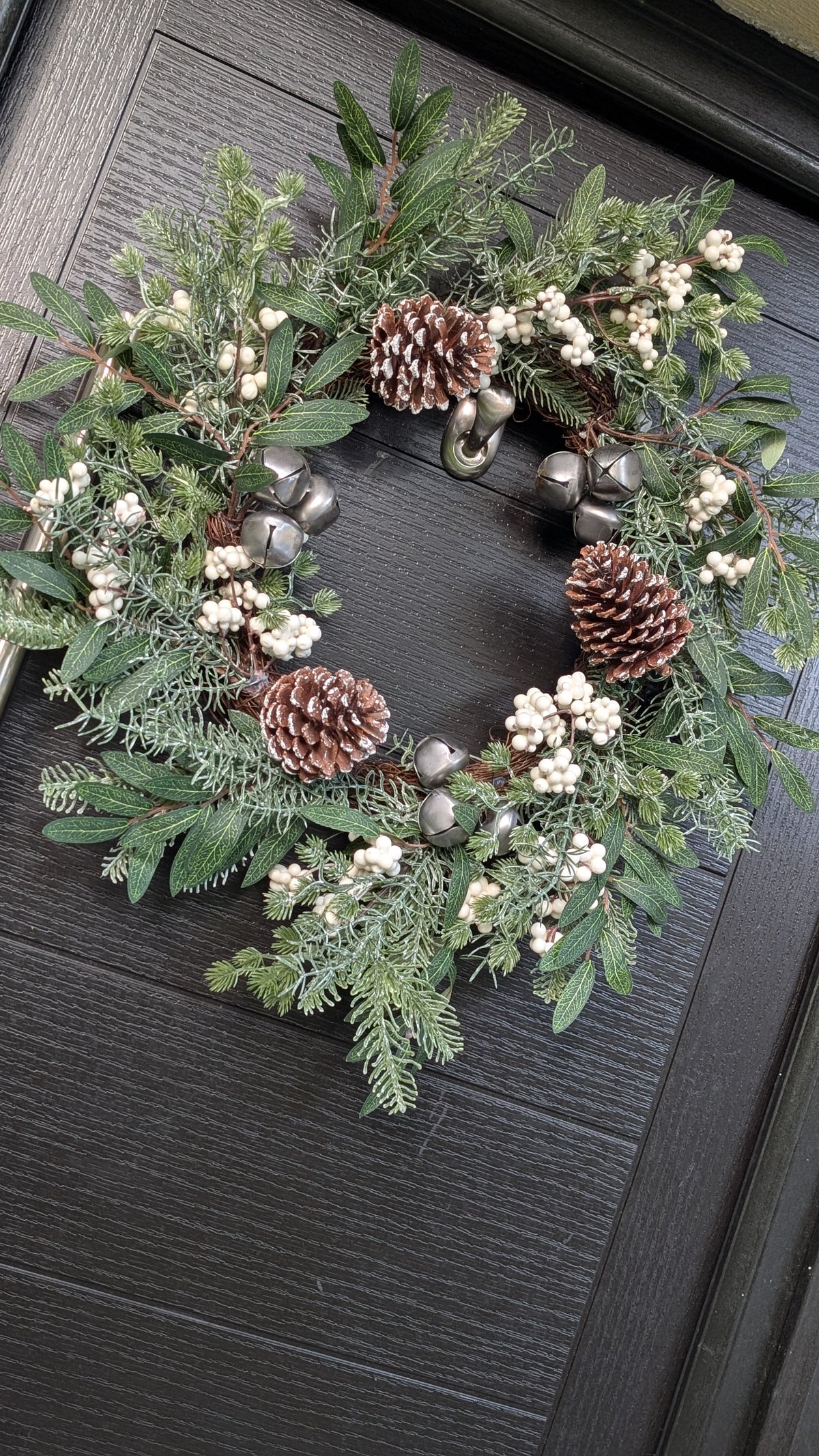 55cm rustic Christmas wreath featuring ivory berries, frosted pinecones, silver bells and mixed green foliage displayed on a dark front door.