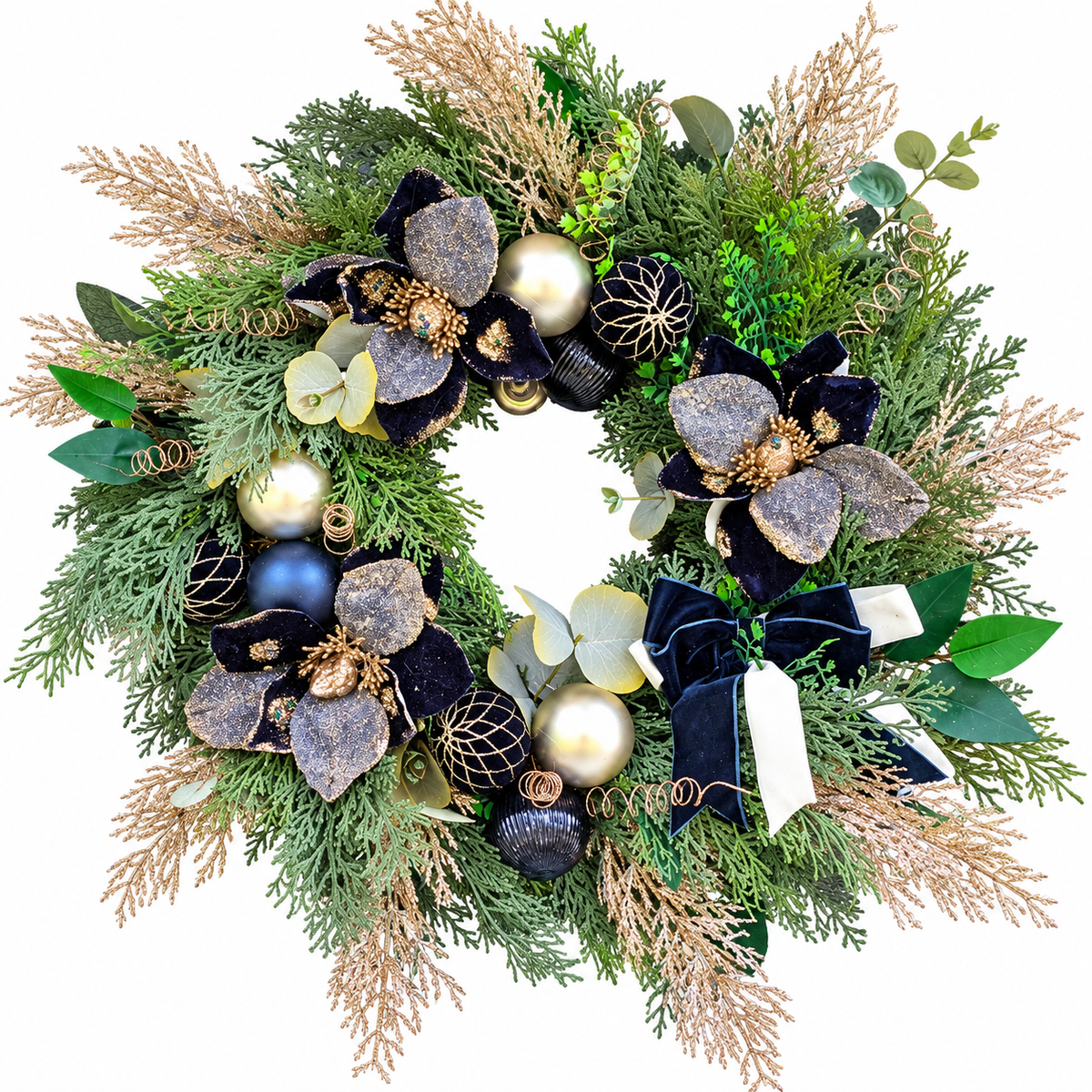A festive holiday wreath decorated with green pine and cedar branches, beige feathery accents, dark blue and metallic gold ornaments, glittery flower petals, and ribbon bows, photographed on a solid white background.