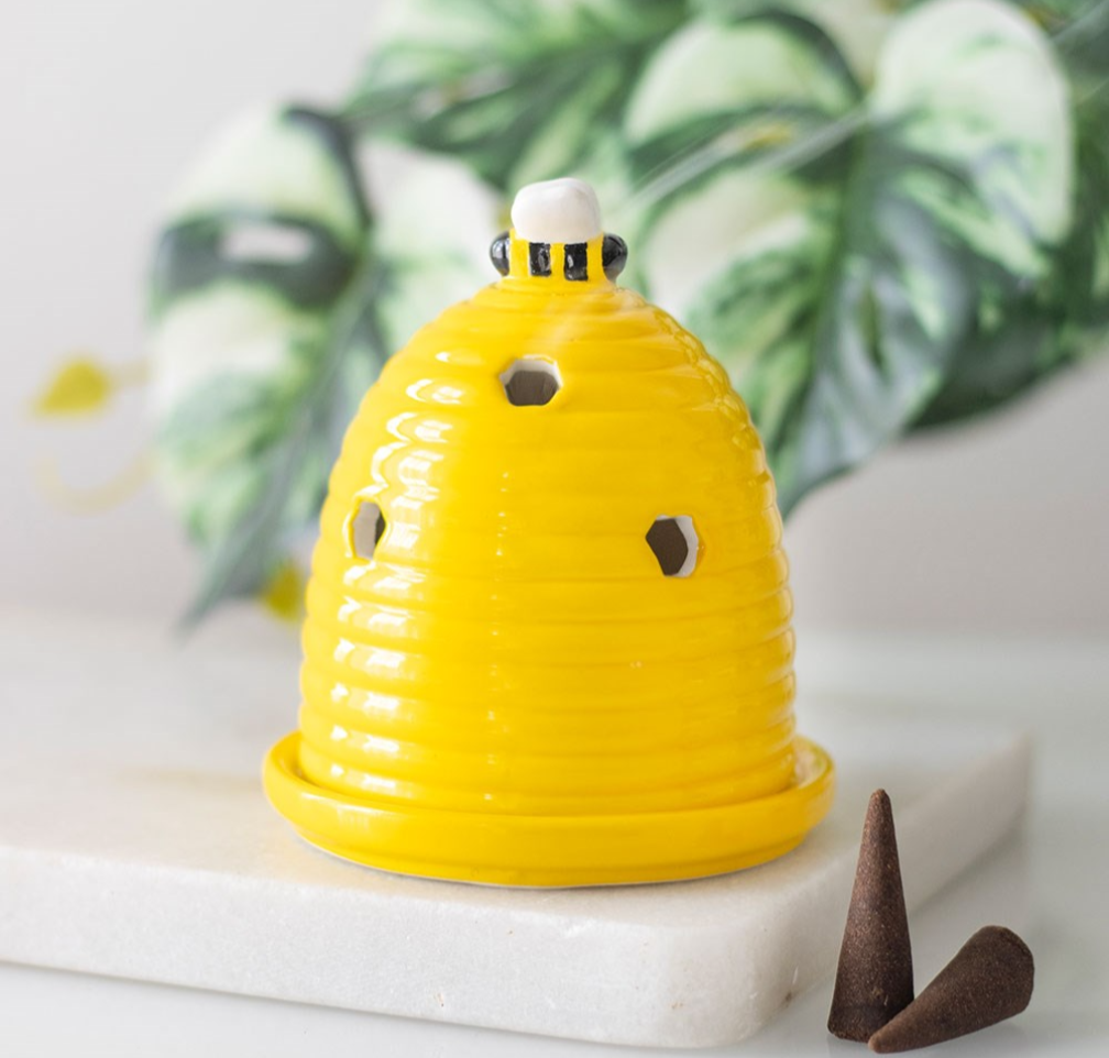 A bright yellow ceramic beehive-shaped incense holder with ventilation holes and a small white top piece, displayed on a white marble slab with green leaves in the background.
