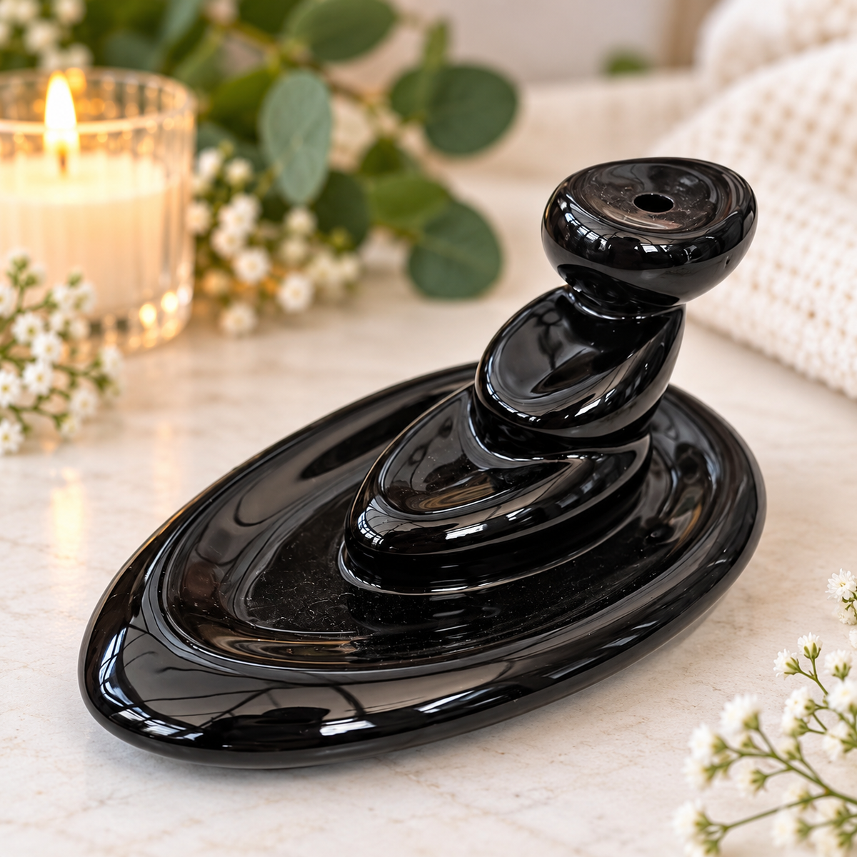 A glossy black ceramic zen stone stack incense burner sits on a light marble surface, surrounded by a lit candle, green leaves, and white flowers.