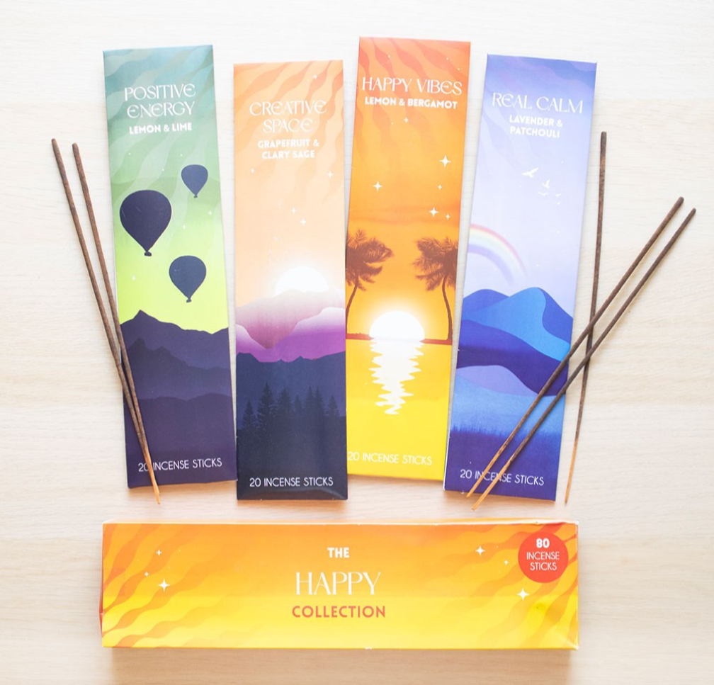 Four colorful incense stick boxes with matching sticks arranged on a light wood surface, plus a larger yellow-orange box labeled 'THE HAPPY COLLECTION' at the bottom. The designs feature different scenic illustrations and text such as 'Positive Energy,' 'Creative Space,' 'Happy Vibes,' and 'Real Calm.