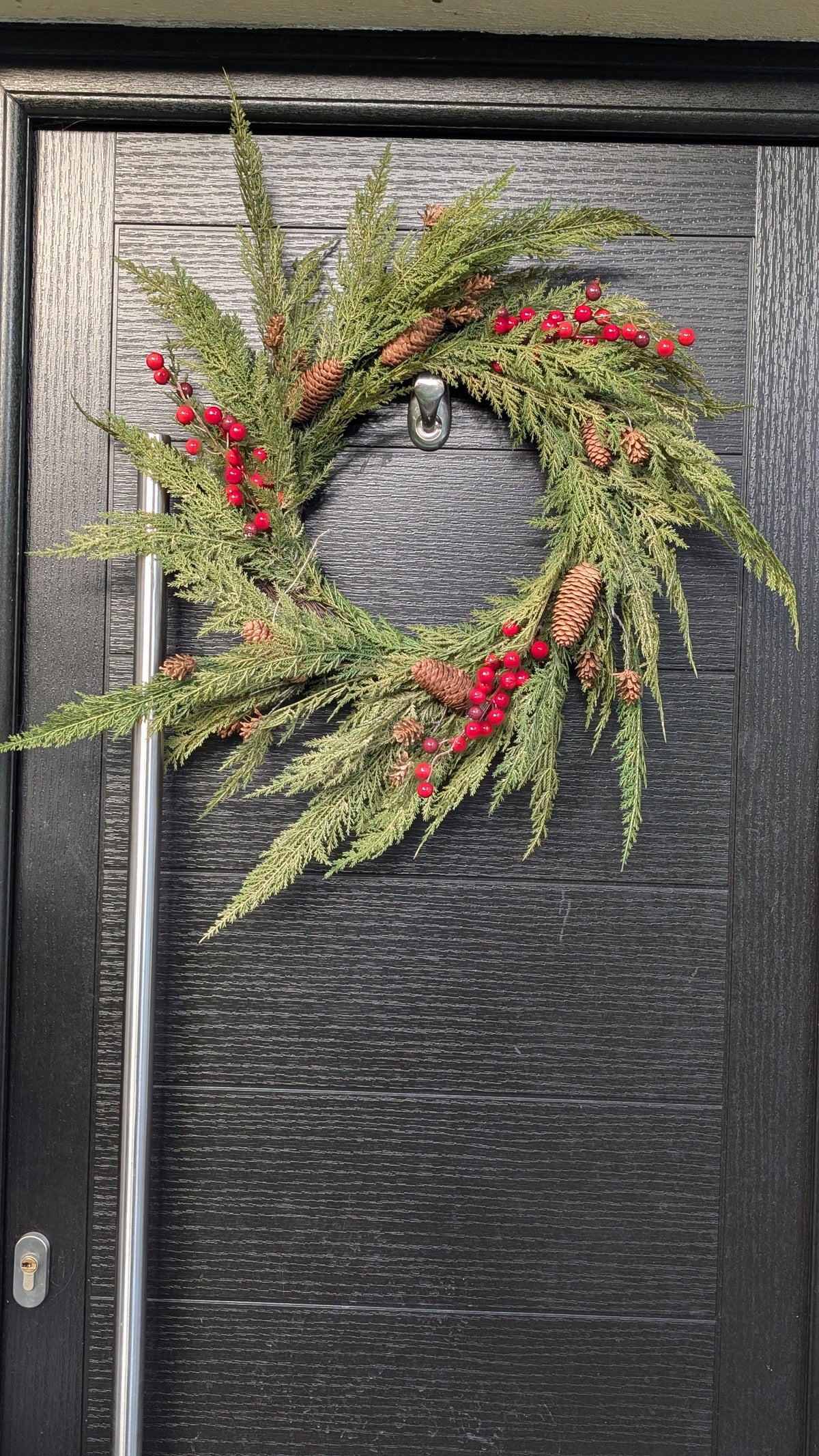 60cm ready-made Cupressus Christmas wreath with cascading green foliage, red berries and natural pinecones displayed on a dark front door.