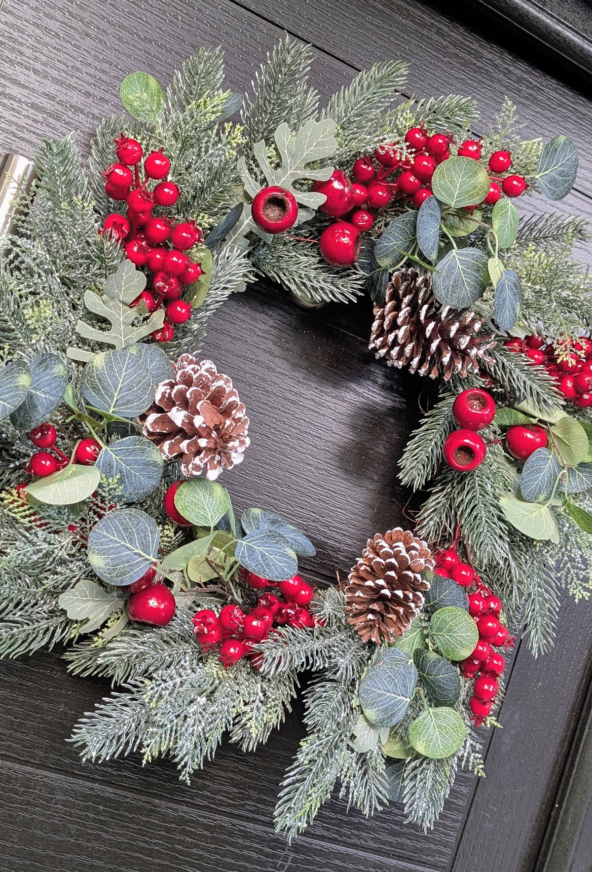 55cm traditional Christmas wreath featuring frosted evergreen foliage, red berries, snowy pinecones and eucalyptus-style leaves against a dark front door.