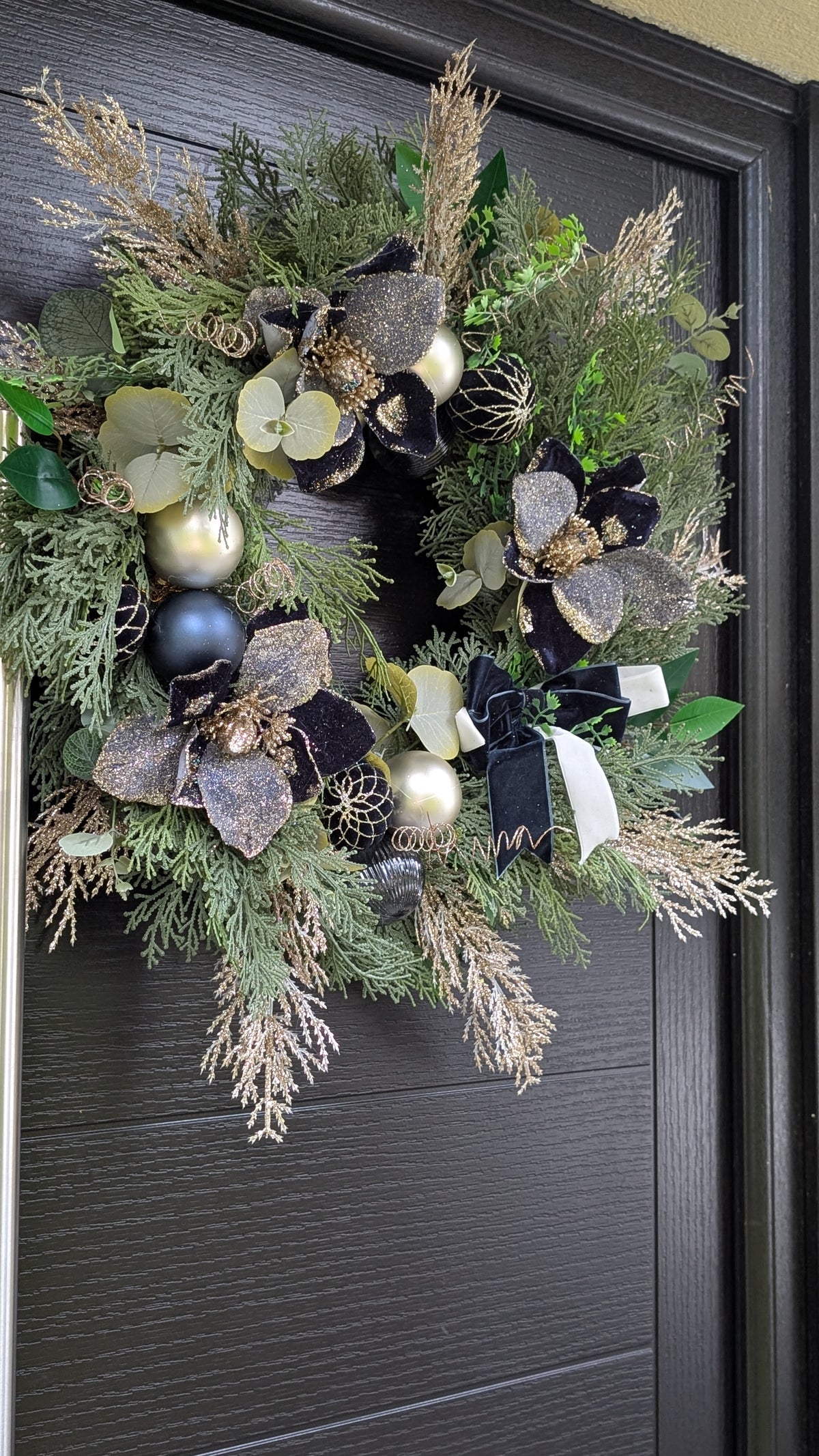 A decorative holiday wreath hanging on a dark charcoal-colored front door. The wreath features green pine and fir branches mixed with dried beige grasses, large black and metallic gold artificial flowers, green eucalyptus leaves, and shiny gold and silver ornaments.