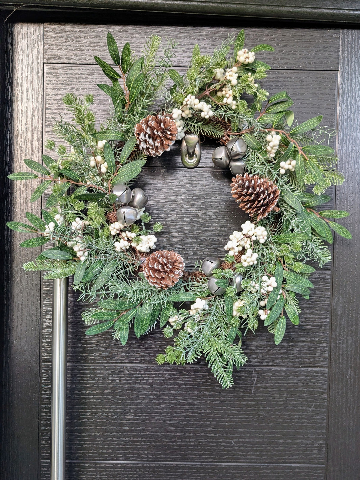 55cm rustic Christmas wreath featuring ivory berries, frosted pinecones, silver bells and mixed green foliage displayed on a dark front door.