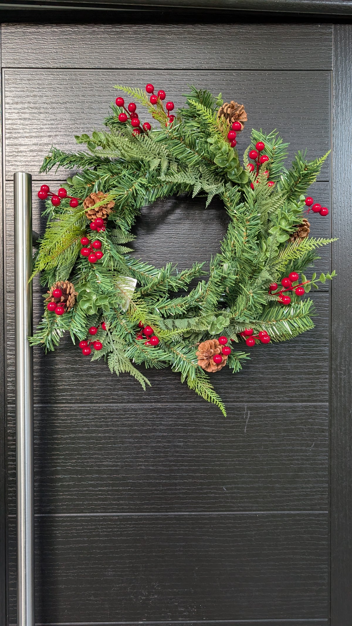 60cm traditional Christmas wreath featuring green spruce foliage, fern-like greenery, bright red berries and natural pinecones displayed on a dark front door.