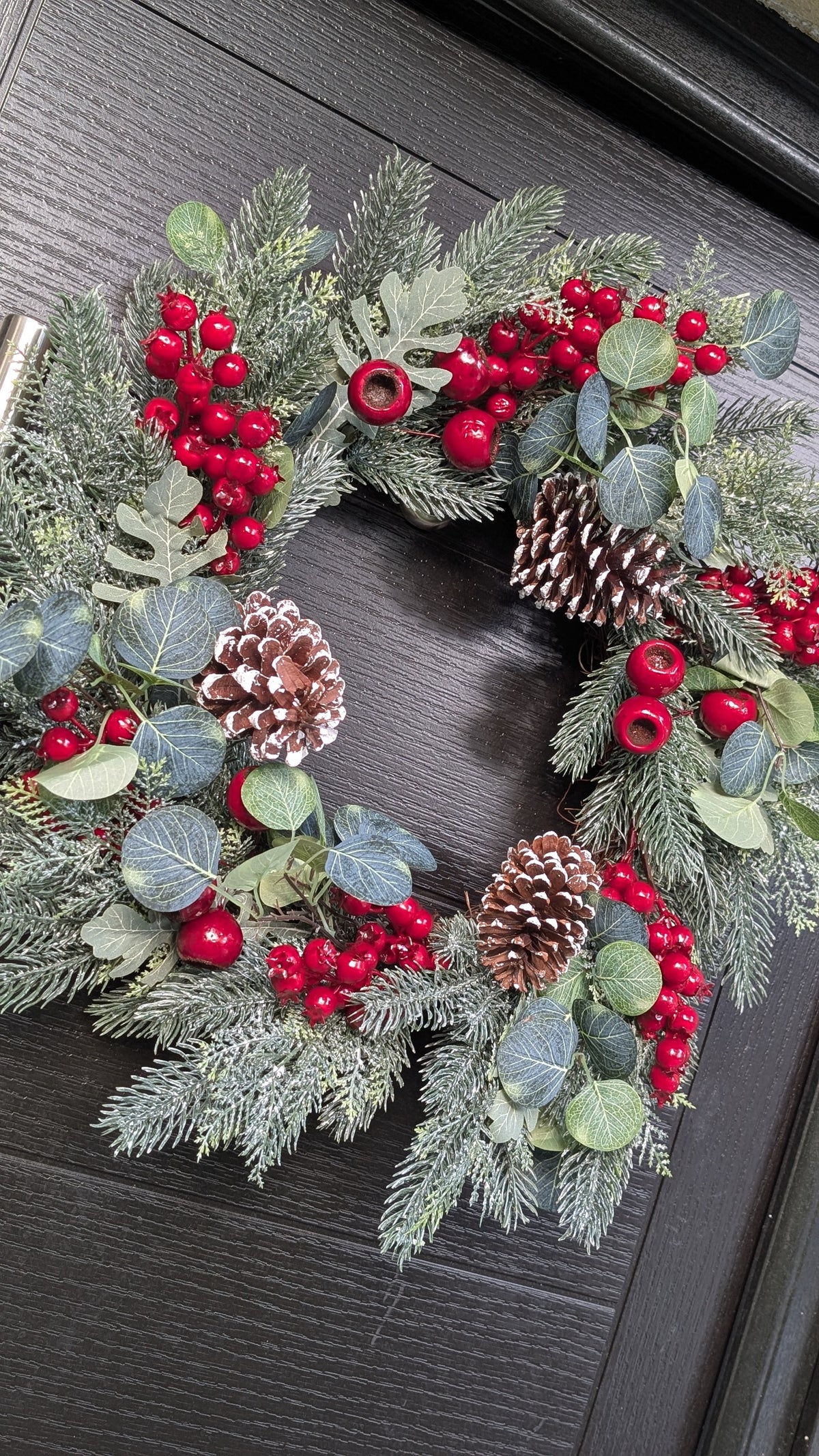 55cm traditional Christmas wreath featuring frosted evergreen foliage, red berries, snowy pinecones and eucalyptus-style leaves against a dark front door.