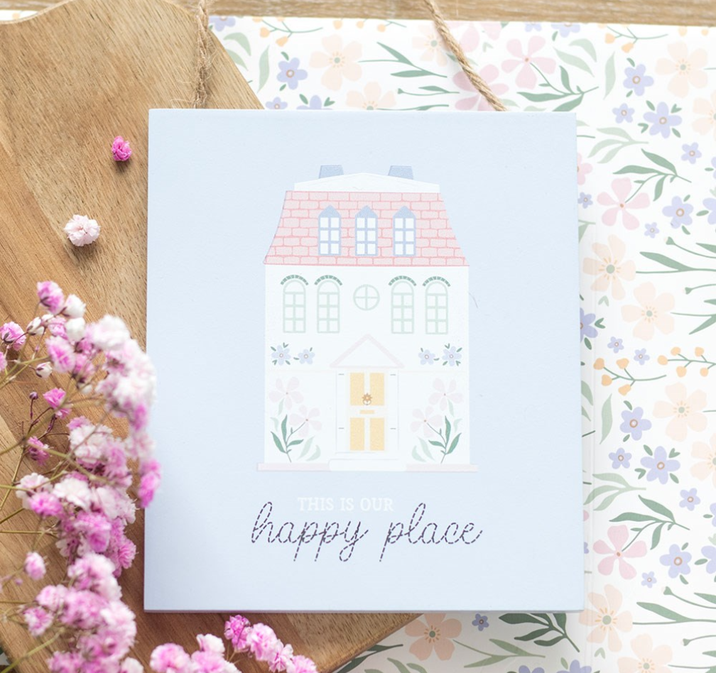 A square light blue greeting card or gift box featuring a cute illustrated house and the words 'this is our happy place,' styled on a wooden surface with floral paper and pink flowers.