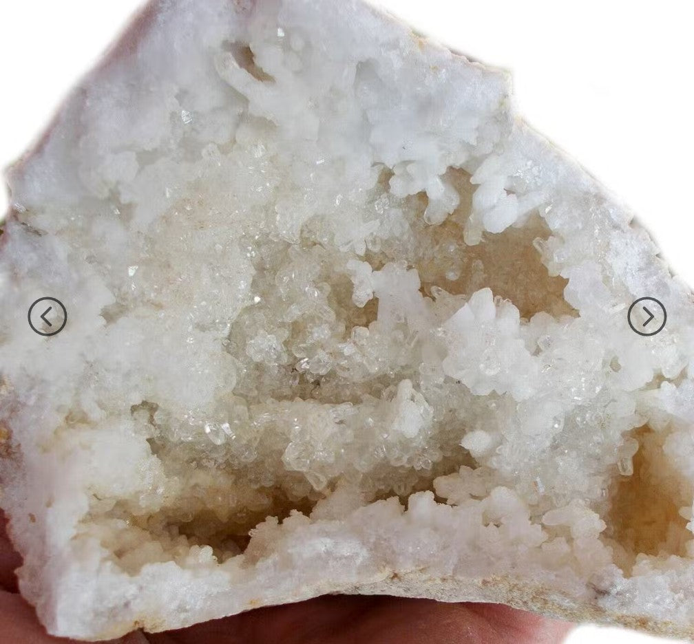A close-up of a white crystal geode or mineral specimen with rough crystalline texture, held against a white background. A hand is partially visible at the bottom edge, and small arrow icons appear on both sides.