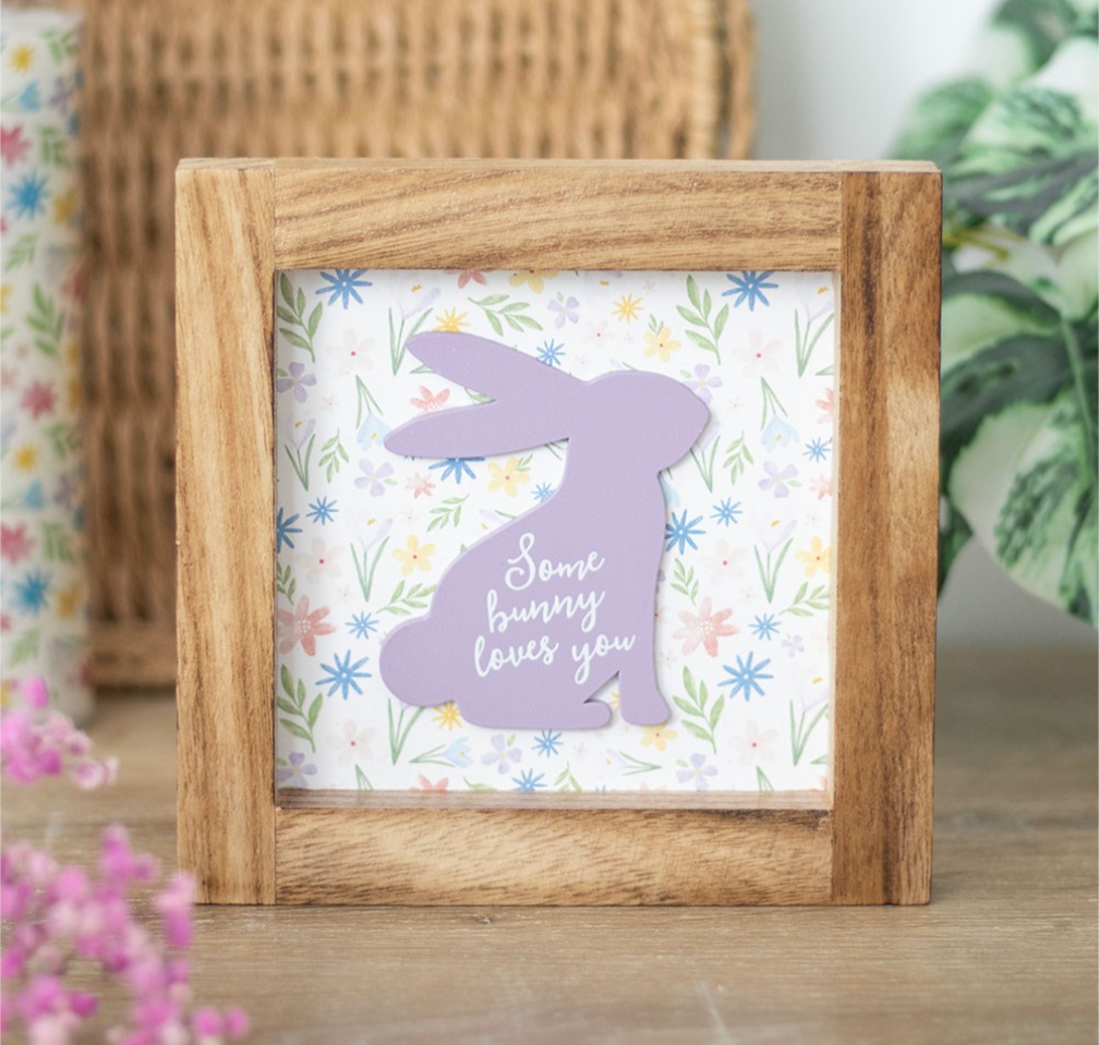 A square wooden framed sign with a lavender bunny silhouette and the phrase 'Some bunny loves you' on a floral background, displayed on a wooden surface with soft-focus decorative plants.