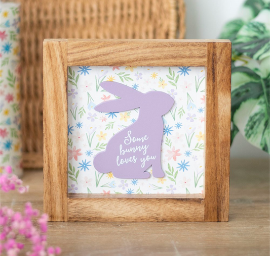 A square wooden framed sign with a lavender bunny silhouette and the phrase 'Some bunny loves you' on a floral background, displayed on a wooden surface with soft-focus decorative plants.
