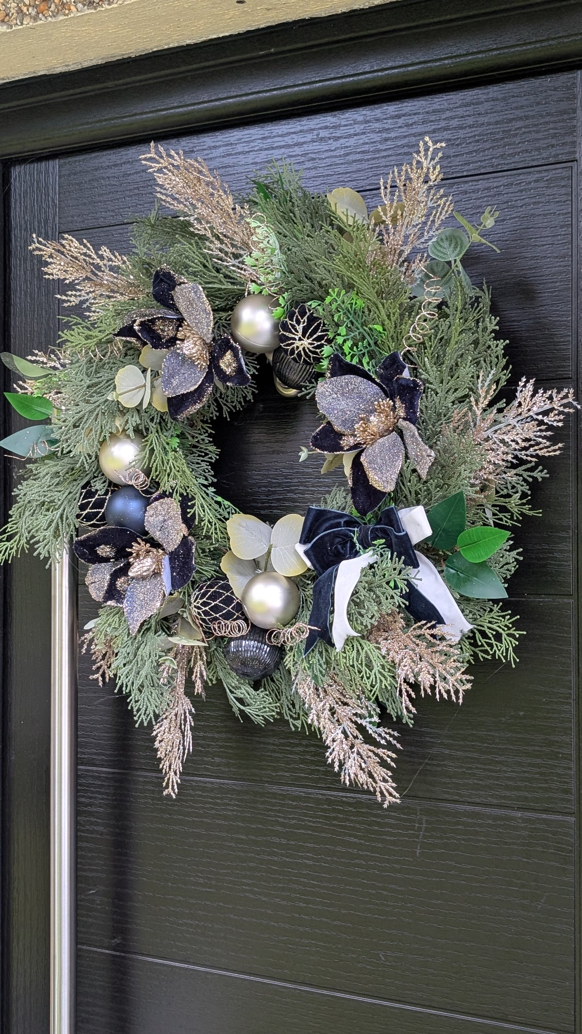 A festive holiday wreath hanging on a dark charcoal-colored front door. The wreath is decorated with green pine and cedar branches, dried beige grasses, large glittery silver and black magnolia flowers, green eucalyptus leaves, and shiny silver and white ornaments.