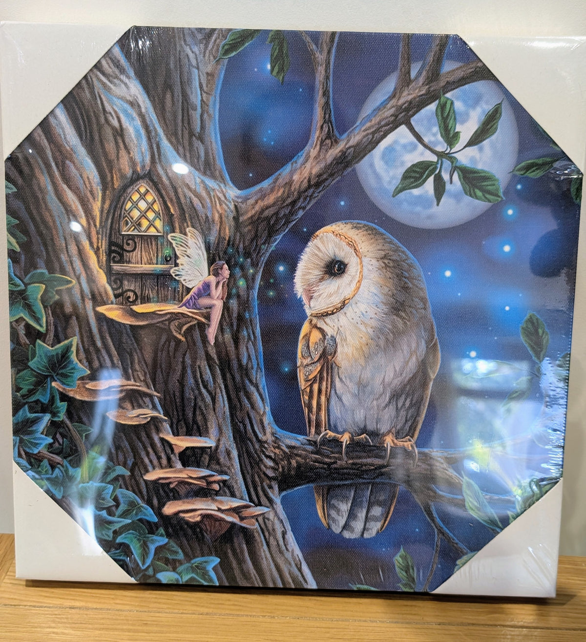 An octagonal decorative plaque or tile featuring a whimsical night scene with a barn owl perched on a tree branch, a small fairy in a treehouse, mushrooms, stars, and a full moon. The item is photographed on a wooden surface with the white backing board visible around the artwork.