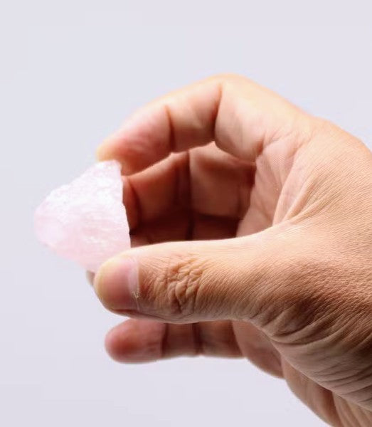 A hand holding a small, rough, pale pink crystal against a plain light gray background.