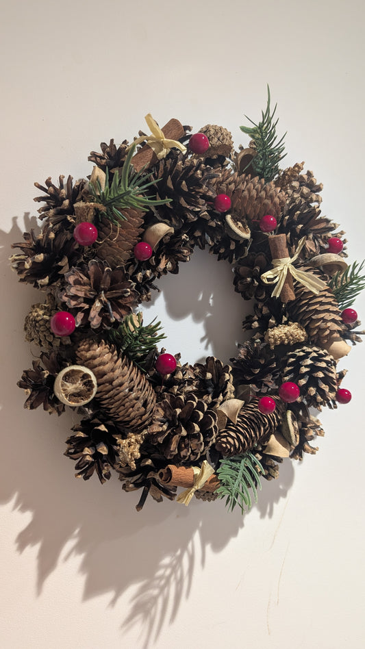 A rustic pinecone wreath decorated with natural brown pinecones, small green pine sprigs, red berries, and cream ribbon bows, hanging against a plain off-white wall.