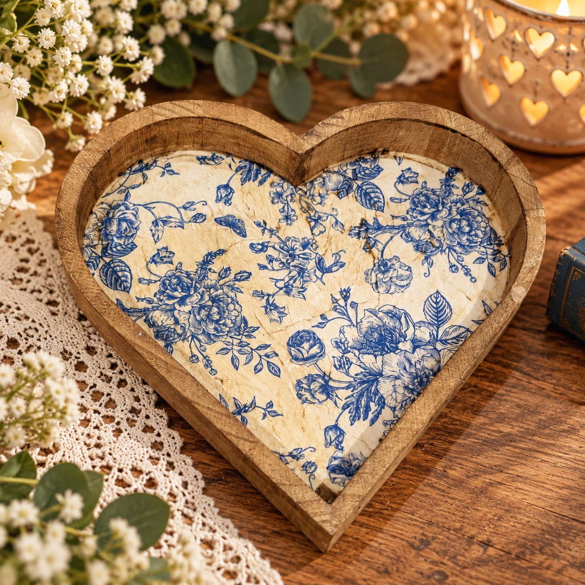 A heart-shaped wooden tray holding a vintage-style cream fabric insert with a blue floral toile pattern, styled on a wooden table with lace, baby's breath, and a candle.