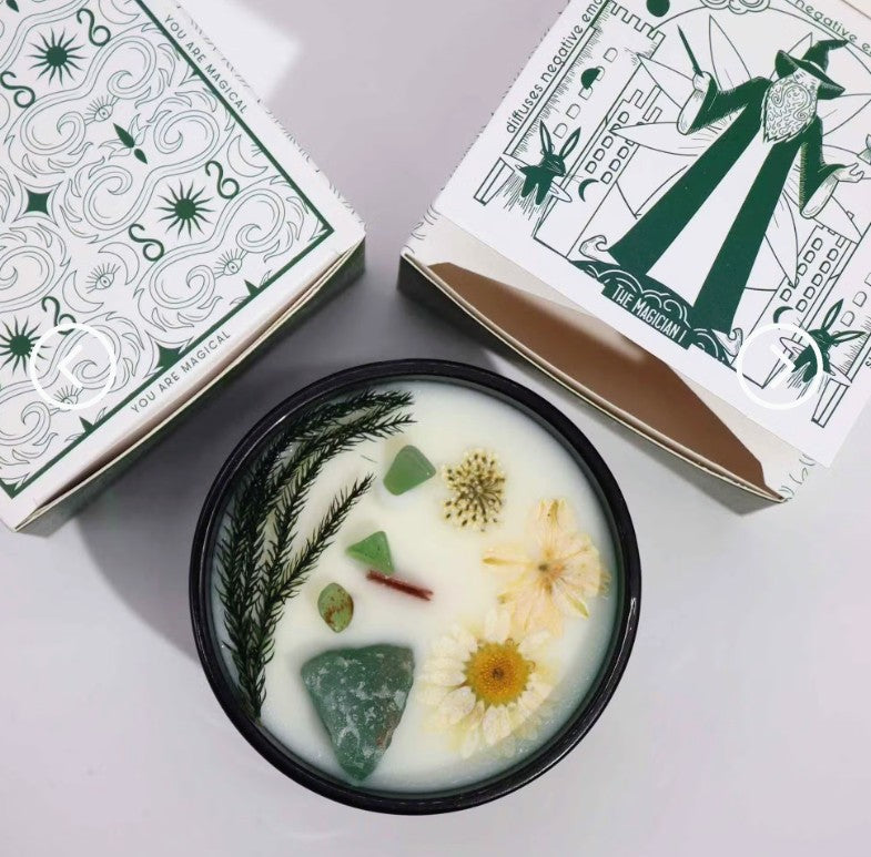 An open black-lidded candle jar filled with white wax and botanical/spell elements, shown with two decorative boxes on a light gray surface. The wax contains green stones, yellow flowers, a dried green herb, and a small red piece.