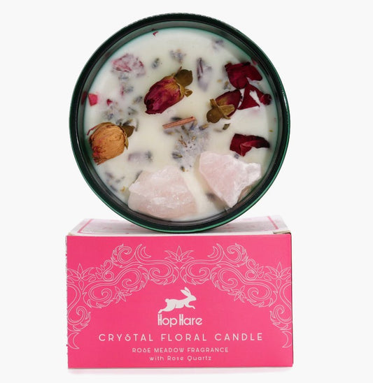A round dark green candle tin filled with white wax and dried botanicals, resting on top of a bright pink box labeled 'hophare CRYSTAL FLORAL CANDLE'.
