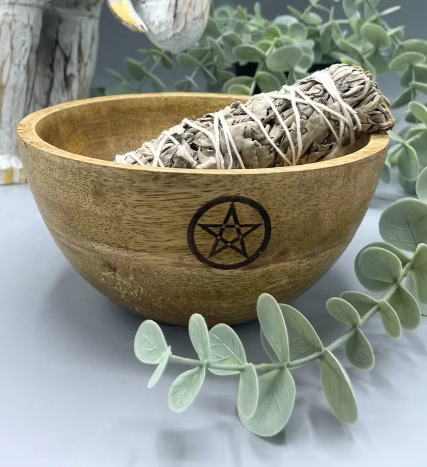 A wooden bowl with a carved pentagram symbol on the front, containing a white sage smudge bundle, styled with eucalyptus leaves on a light gray surface.