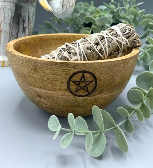 A wooden bowl with a carved pentagram symbol on the front, containing a white sage smudge bundle, styled with eucalyptus leaves on a light gray surface.