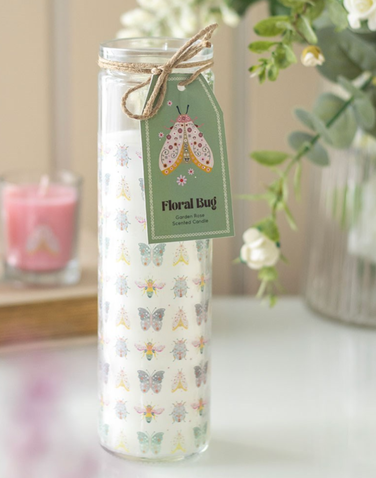 A tall glass jar candle with a white wax body printed with colorful butterfly and moth motifs, tied with a twine bow and a green paper tag reading 'Floral Bug.' The candle is photographed on a tabletop with soft-focus flowers and another pink candle in the background.