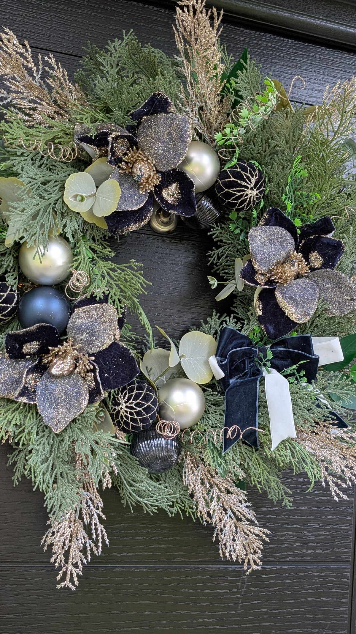 A festive Christmas wreath made of green pine and cedar branches, decorated with black and silver glittery flowers, velvet ribbons, gold pinecones, and metallic baubles, hanging on a dark wood door.