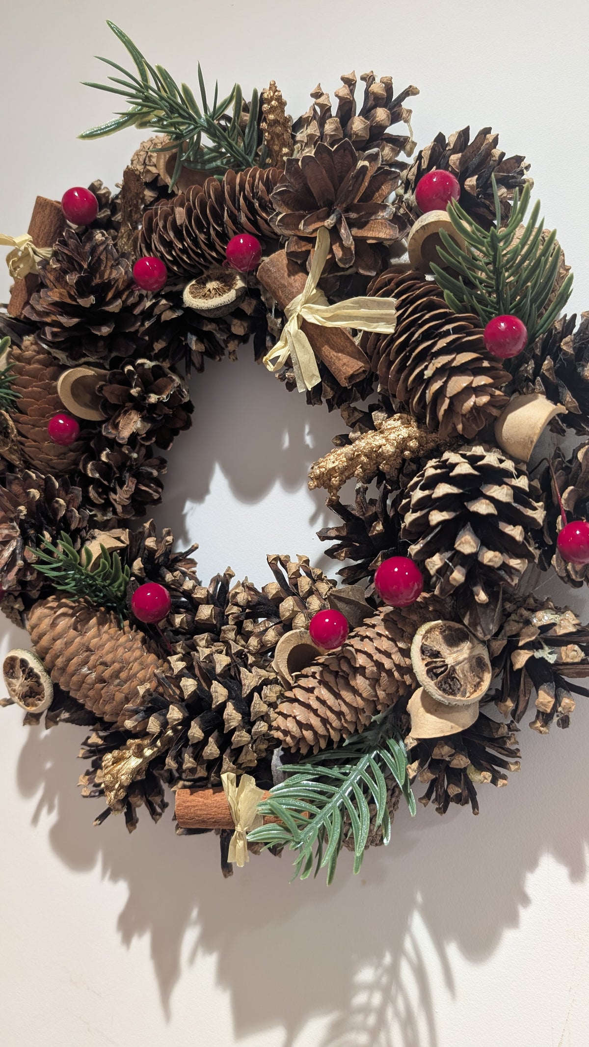 A rustic pinecone wreath decorated with natural brown pinecones, green pine needles, small red berries, dried citrus slices, cinnamon sticks, and a cream ribbon bow, shown against a plain off-white background.