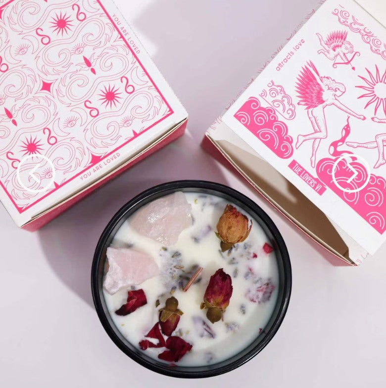 A black bowl filled with a white wax candle and botanical pieces, styled with two decorative pink and white gift boxes on a pale pink background.