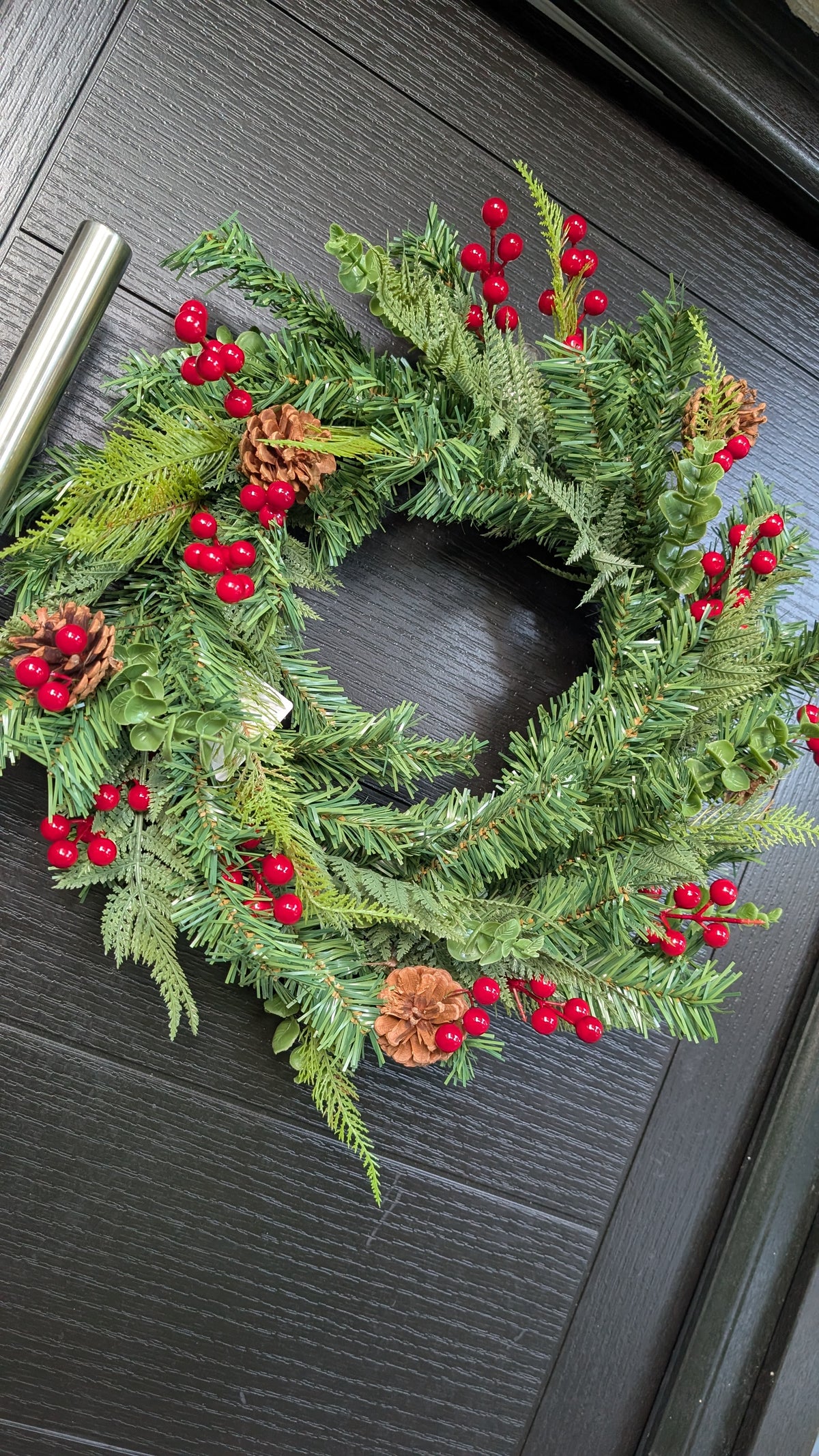 60cm traditional Christmas wreath featuring green spruce foliage, fern-like greenery, bright red berries and natural pinecones displayed on a dark front door.
