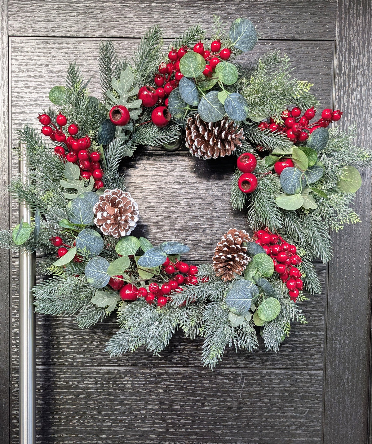 55cm traditional Christmas wreath featuring frosted evergreen foliage, red berries, snowy pinecones and eucalyptus-style leaves against a dark front door.