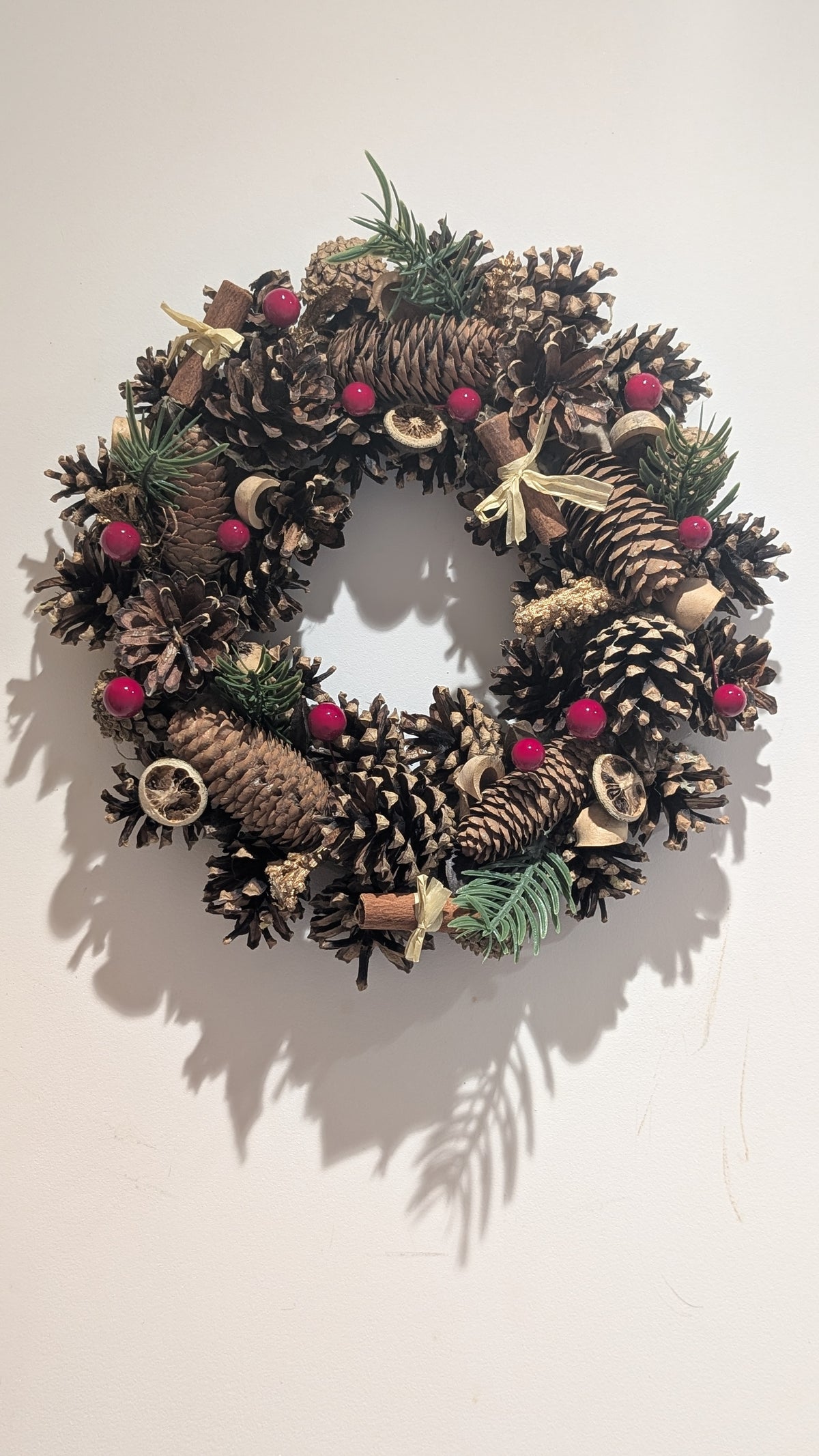 A rustic pinecone wreath decorated with natural brown pinecones, small green pine sprigs, dried red berries, and light beige ribbon accents, hanging against a plain off-white wall.