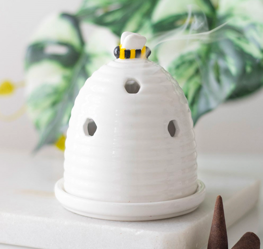 A white ceramic beehive-shaped incense burner with ventilation holes, a small bee topper with yellow and black stripes, and visible rising smoke, displayed on a white stone surface with blurred green leaves in the background.