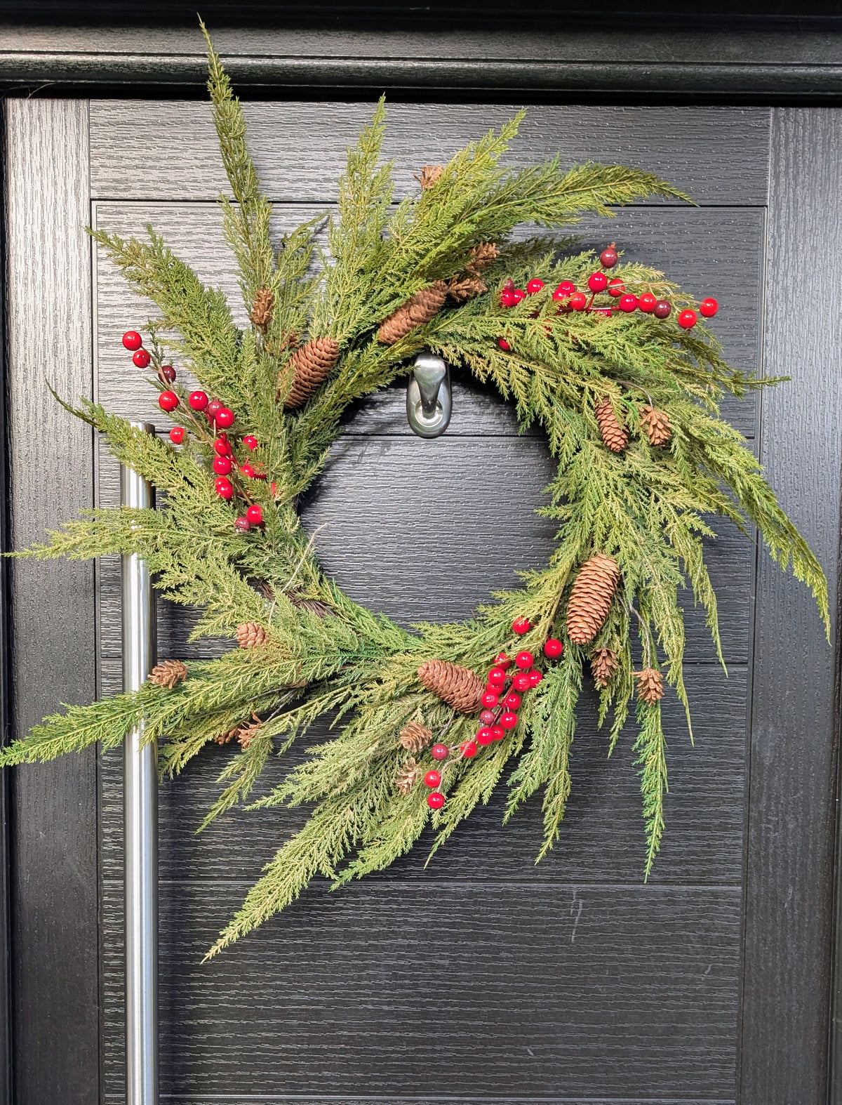 60cm ready-made Cupressus Christmas wreath with cascading green foliage, red berries and natural pinecones displayed on a dark front door.